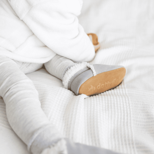 Baby auf dem Bett liegend, mit hellgrauen Dotty Fish Barfußpantoffeln aus Wildleder, mit Fleece gefüttert.