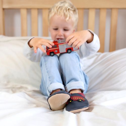 Kleiner Junge sitzt auf dem Bett, trägt marineblaue Dotty Fish-Schuhe mit rotem Feuerwehraufdruck und spielt mit einem Spielzeug-Feuerwehrauto.