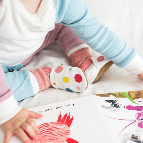 Toddler girl reading a book while wearing white leather baby shoes with colourful spotty design from Dotty Fish