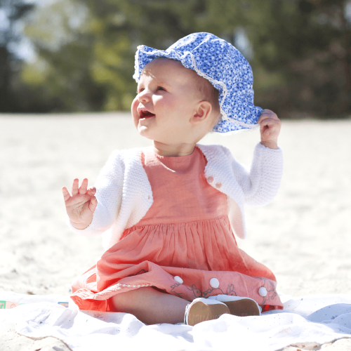 Kleinkind Mädchen sitzt am Strand und trägt weiße Leder Dotty Fish erste Walker barfuß Sandalen mit weißen Blumen.