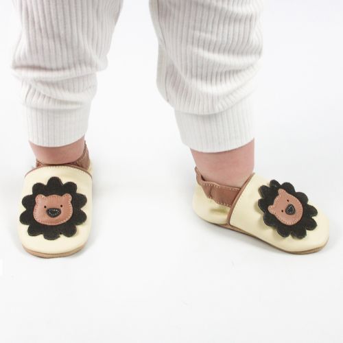 Baby trägt cremefarbene Dotty Fish-Schuhe mit Löwenmotiv und weißem Babywuchs.