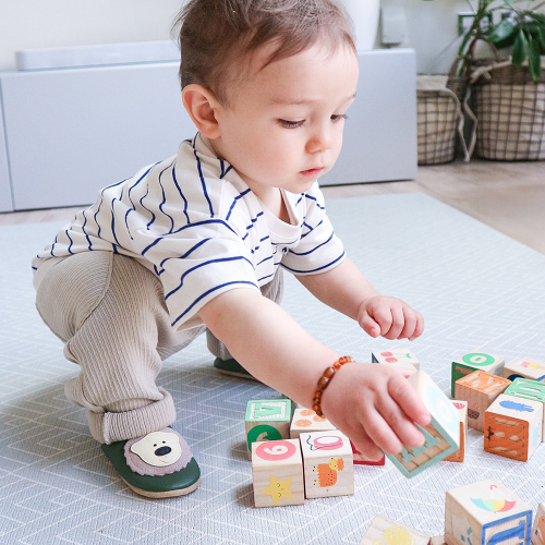 Ein kleiner Junge sitzt auf dem Boden, trägt grüne Dotty Fish Schuhe mit cremefarbenem Bärenmuster und spielt mit Holzklötzen.