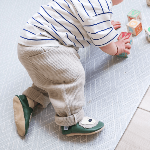 Ein kleiner Junge sitzt auf dem Boden und trägt grüne Dotty Fish Schuhe mit cremefarbenem Bärenmuster.