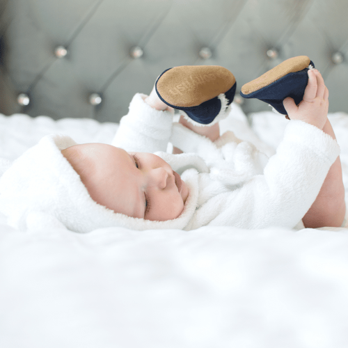 Das Baby liegt auf dem Bett und trägt marineblaue Dotty Fish Barfußpantoffeln aus Wildleder mit Fleecefutter.