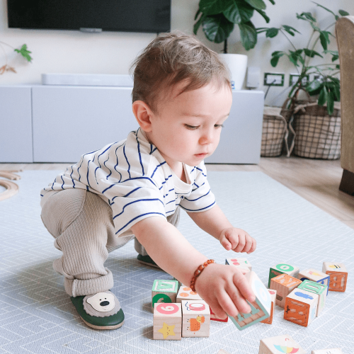 Ein kleiner Junge sitzt auf dem Boden, trägt grüne Dotty Fish Schuhe mit cremefarbenem Bärenmuster und spielt mit Holzklötzen.