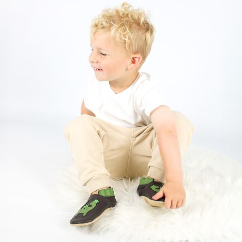 Ein Kleinkind sitzt auf dem Boden und trägt braune Dotty Fish Schuhe mit grünem Dinosaurier-Muster.
