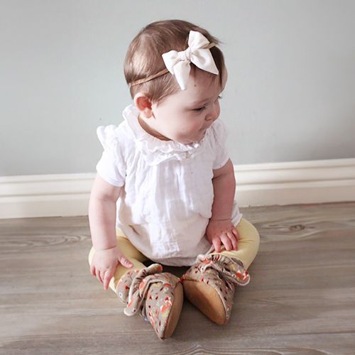 Baby-Mädchen sitzt und trägt hellbraune Dotty Fish-Schuhe aus Baumwolle mit weißem Fleece-Futter und Waldtiermuster.