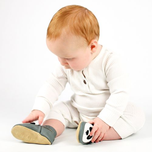 Kleiner Junge sitzt auf dem Boden, trägt graue Dotty-Fish-Schuhe mit weißem und schwarzem Panda-Muster und schaut auf seine Füße.