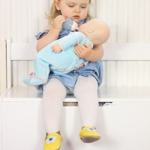 Little girl playing with doll while wearing yellow daisy soft leather baby shoes from Dotty Fish 