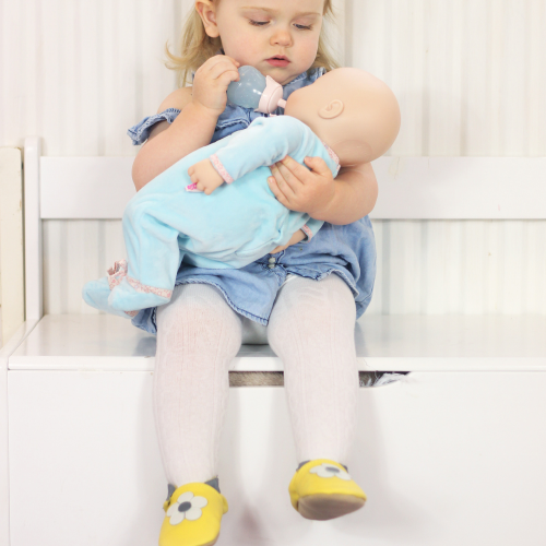 Mädchen trägt gelbe Dotty Fish Lauflernschuhe aus Leder mit weißer Blume und spielt mit einer Puppe.