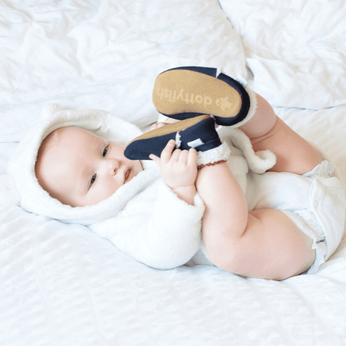 Das Baby liegt auf dem Bett und trägt marineblaue Dotty Fish Barfußpantoffeln aus Wildleder mit Fleecefutter.