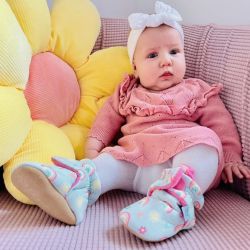 Baby-Mädchen in blassblauen Dotty-Fish-Stiefeln aus Baumwolle mit rosa Fleece-Futter und Regenbogenmuster, sitzend mit Blumenkissen.